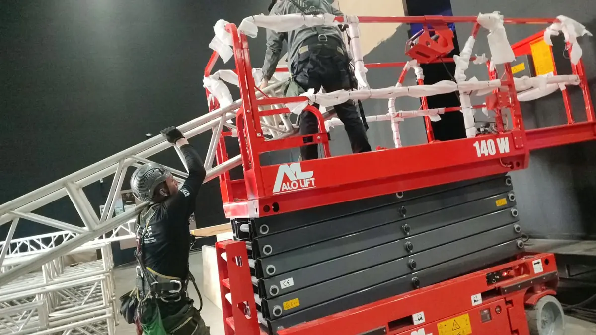 Tijera eléctrica y técnicos con arnés montando una estructura de truss